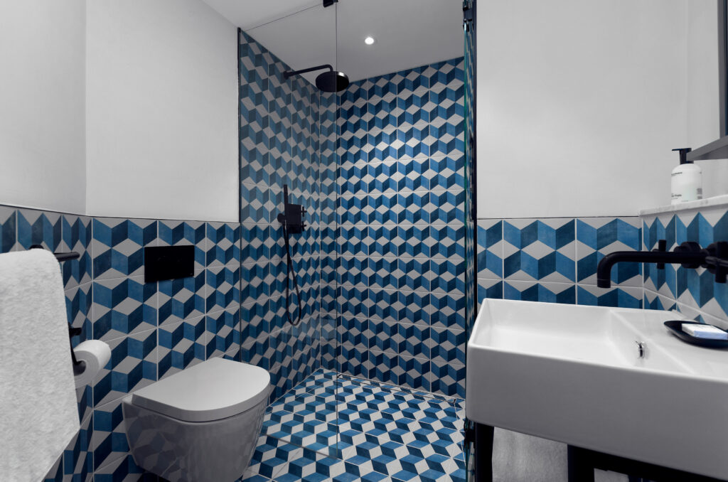 City Single en-suite at Kimpton de Witt with walk-in shower behind glass screen, wall-hung toilet, wall-mounted sink and matt black fittings, blue cube-pattern cement tile to walls and floor.