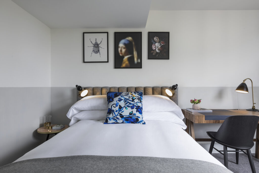 City Single room at Kimpton de Witt with single bed and padded headboard beneath three framed artworks, reading lamps either side, desk with chair to one side and dark wood floor with patterned rug.