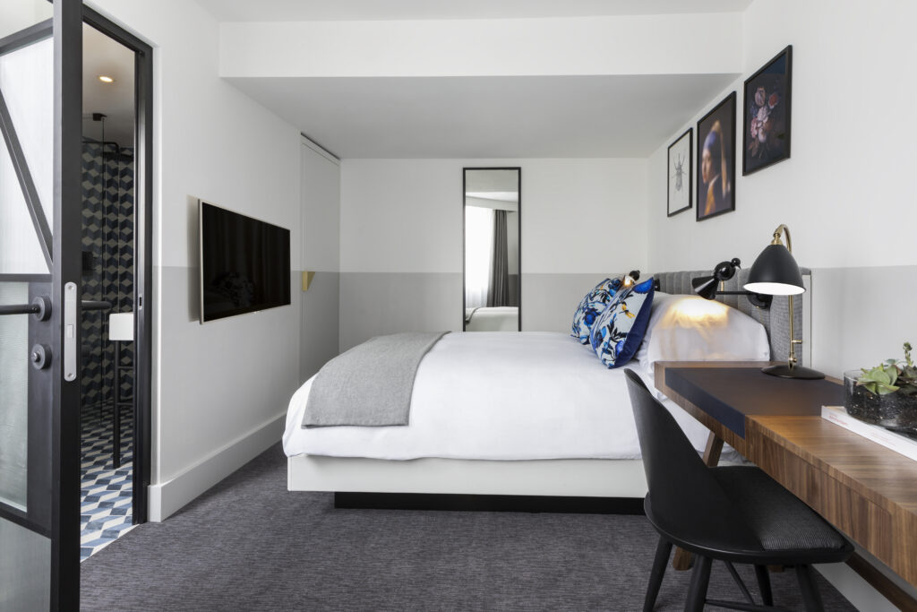 City Single room view at Kimpton de Witt showing wall-mounted TV opposite the bed and full-length mirror, with en-suite bathroom visible through doorway.