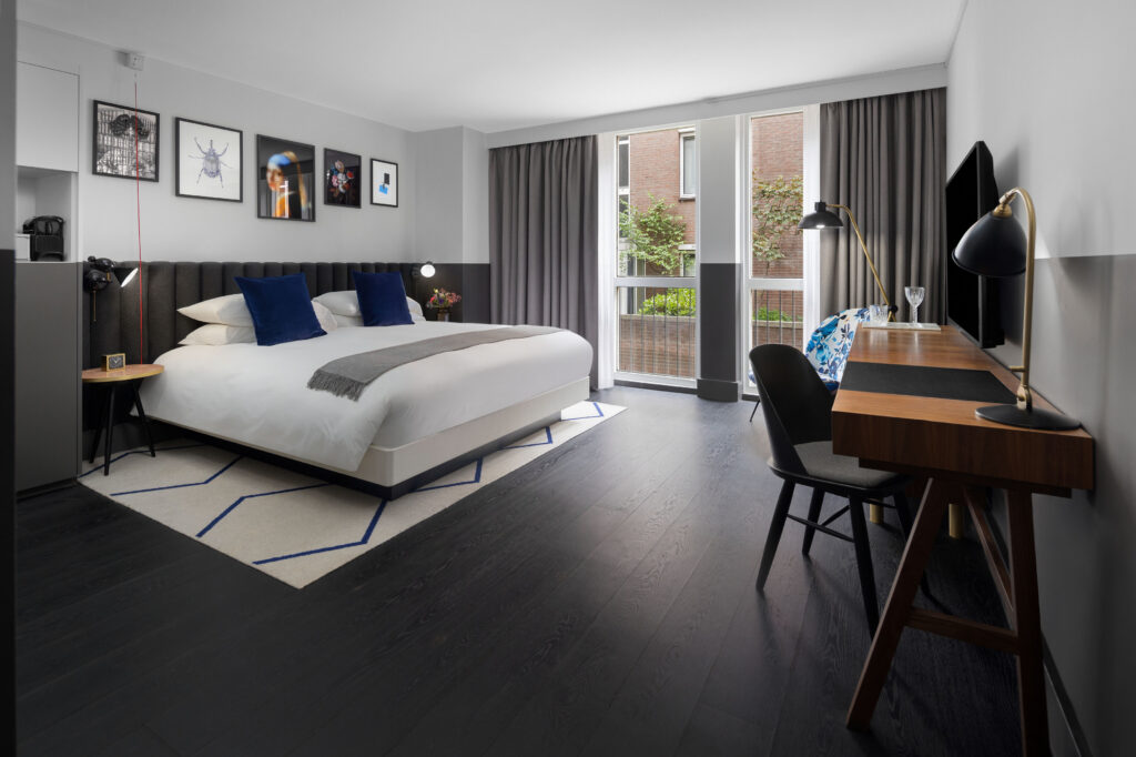 King Bed Premium Accessible room at Kimpton de Witt with king bed beneath a gallery wall of five framed artworks, desk and chair to one side, dark wood floor with patterned rug and large window with full curtains.