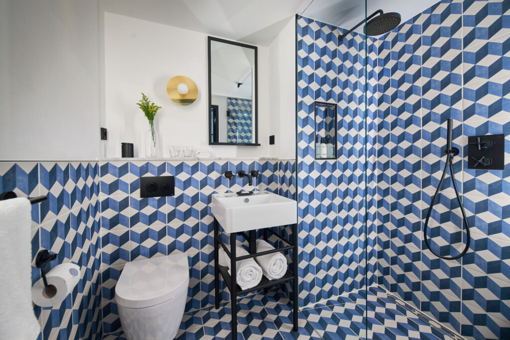 King Bed Premium with Terrace en-suite at Kimpton de Witt with walk-in shower behind glass screen, wall-hung toilet, wall-mounted sink and matt black fittings, blue cube-pattern cement tile throughout.