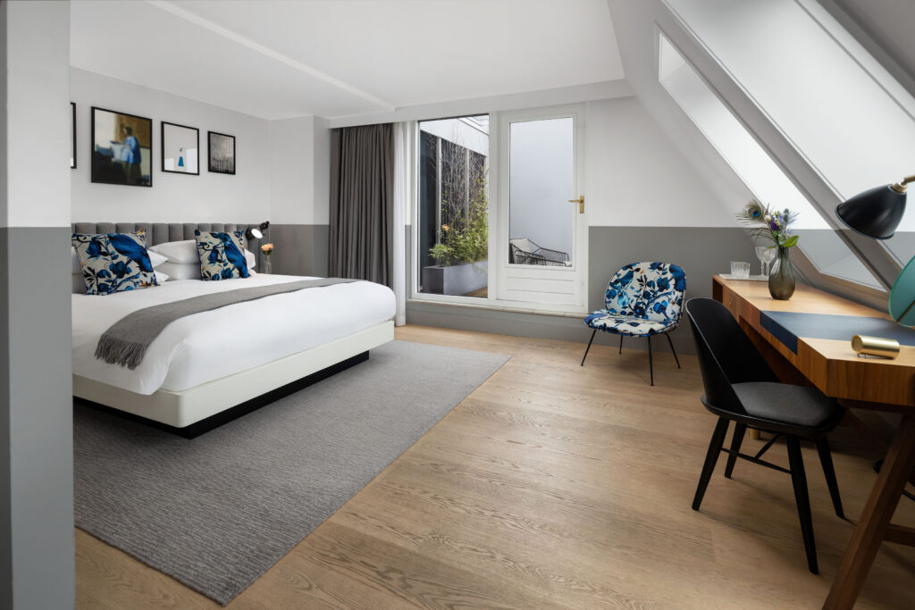 King Bed Premium with Terrace at Kimpton de Witt with sloped loft ceiling and skylight, king bed beneath framed prints, French doors to private terrace and desk to one side.