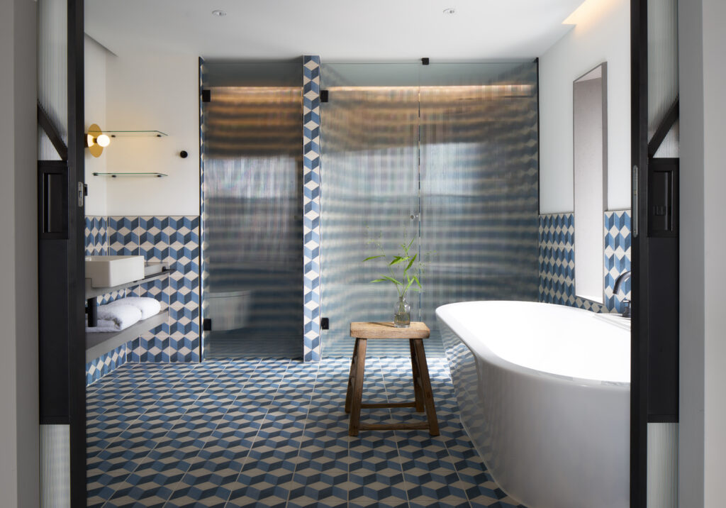 Penthouse de Witt bathroom showing walk-in shower behind fluted glass screens and freestanding bathtub to one side, separate vanity area with glass shelving and blue cube-pattern tile.