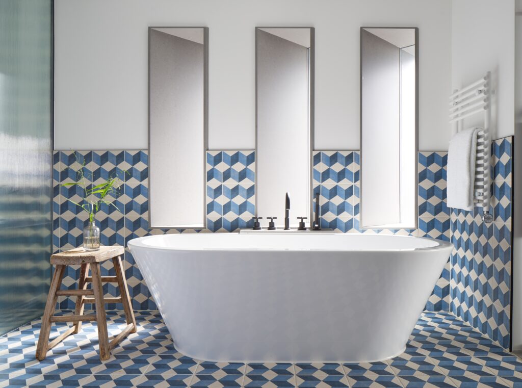 Penthouse de Witt bathroom featuring freestanding bathtub set beneath three tall mirror niches, wall-mounted black tap with hand shower attachment, towel radiator and blue cube-pattern cement tile.