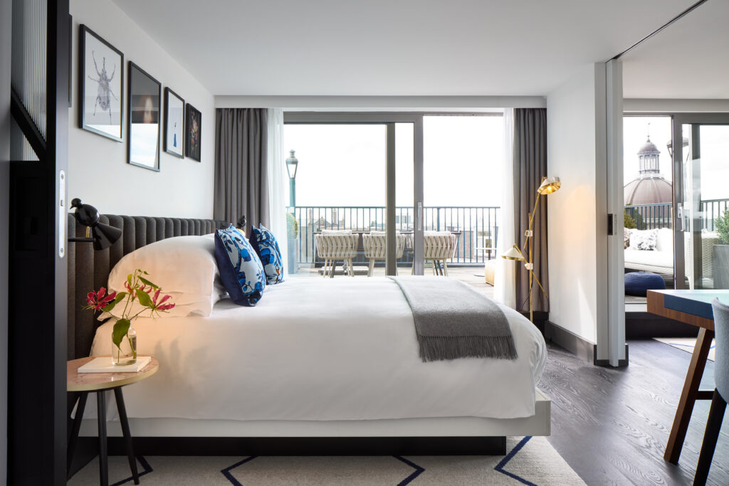 Penthouse de Witt bedroom with king bed beneath framed artwork, sliding doors to the private terrace with dining setting and view to historic Amsterdam dome architecture.