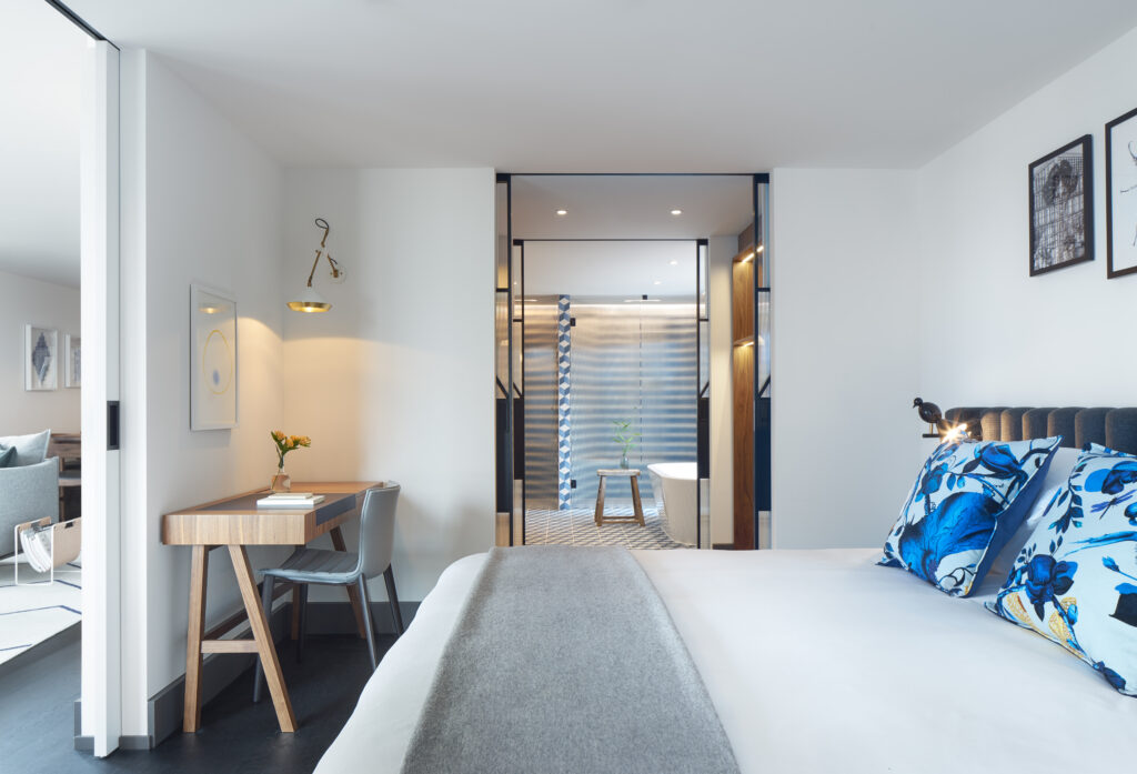 Penthouse de Witt bedroom view from corridor entrance with king bed centre frame, desk with chair to one side and en-suite bathroom with freestanding tub visible beyond.