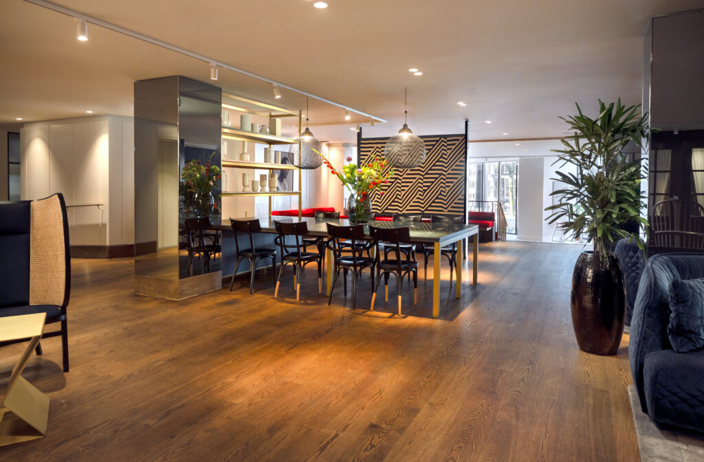 Kimpton de Witt public lounge with long communal dining table beneath mesh pendant lights, brass display shelving alongside, geometric wood feature wall and cane and velvet armchair seating to the side