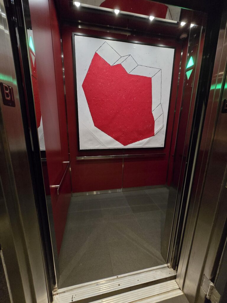 Guest lift interior at Hotel Park Ave NYC. Red-panelled cab with framed artwork, handrail, and a Braille floor plate at the door. Doors open at a level threshold.