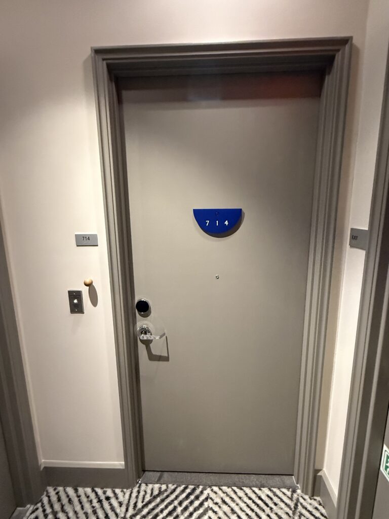 Accessible bedroom 714 entry door from the corridor at Hotel Park Ave. Blue half-disc room number plaque at viewing height. Wall-mounted room number plate to the left of the door at lower height. Lever handle and electronic lock. Patterned corridor carpet meets a flush threshold. Exit signage to the right.