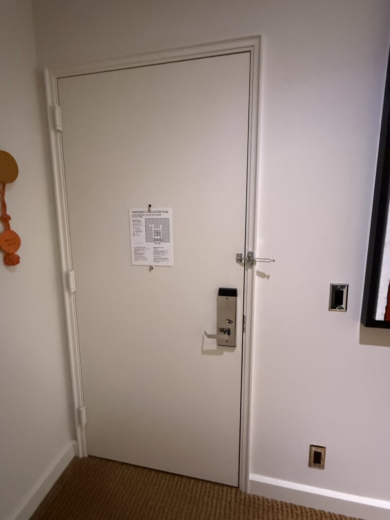 Accessible bedroom entry door from inside the room at Hotel Park Ave. Lever handle with thumb-turn deadbolt. Door-mounted security latch above the handle. Evacuation plan posted on the door. Flush threshold between corridor tile and bedroom carpet. Light switches and key fob recess on the wall to the right.