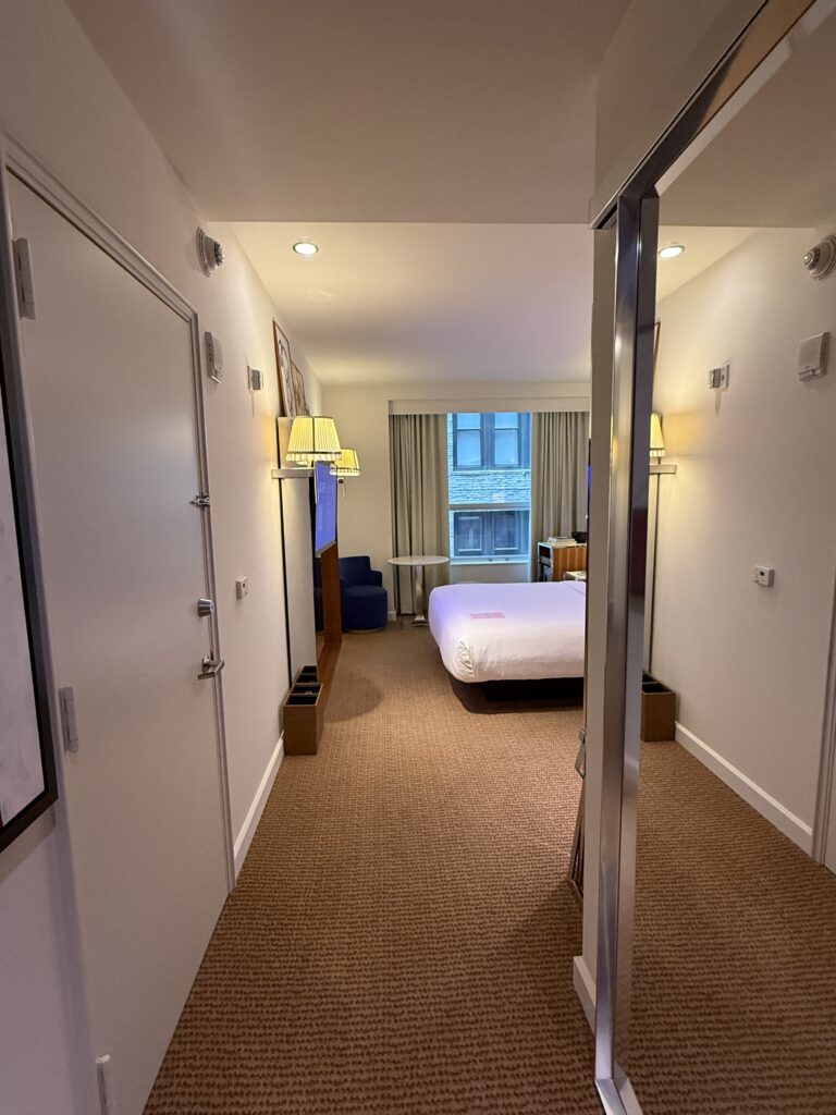 Entry hallway inside the accessible bedroom at Hotel Park Ave. Carpeted floor leads from the entrance through to the bed at the end of the room. Full-height mirrored wardrobe doors on the right. Wall sconces with pleated shades. Clear circulation width along the hallway.