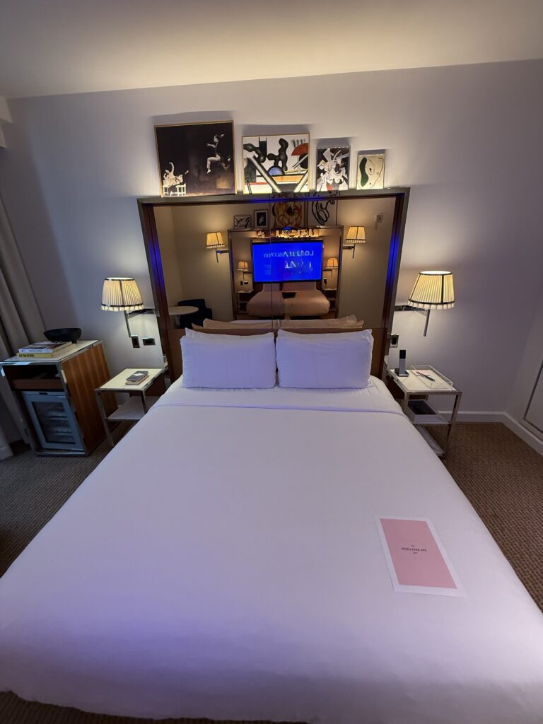 Accessible bedroom at Hotel Park Ave. King bed with white linens centred under a mirrored bronze headboard. Clear floor space along both sides of the bed. Pleated wall sconces and bedside tables with lamps. TV mounted within the headboard. Refrigerator unit at the left bedside. Framed artwork displayed above the headboard.