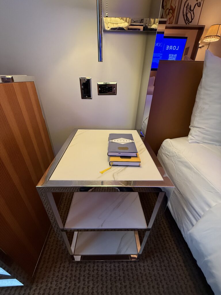 Left bedside table in the accessible bedroom at Hotel Park Ave. Marble-topped chrome two-tier side table beside the bed. Books stacked on the surface. Mains and USB outlets on the wall above the table at reachable height. Pleated lamp arm above.
