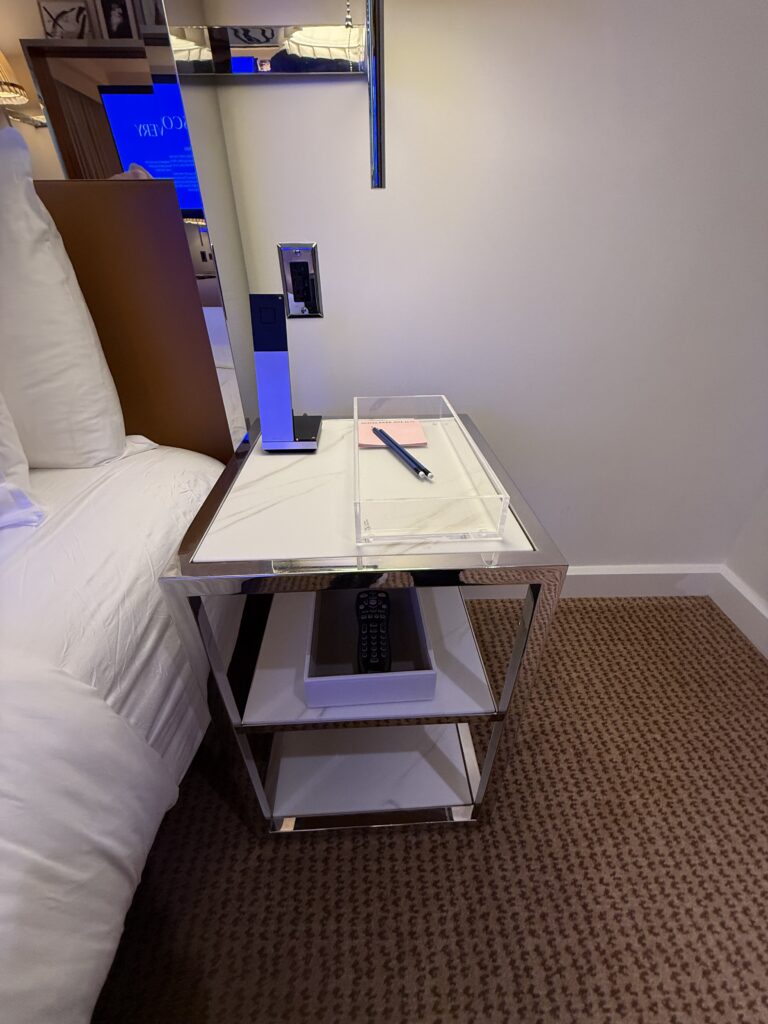 Right bedside table in the accessible bedroom at Hotel Park Ave. Marble-topped chrome two-tier side table. Cordless telephone and notepad on the surface. TV remote on the lower shelf. Mains outlet with USB ports on the wall above at reachable height.