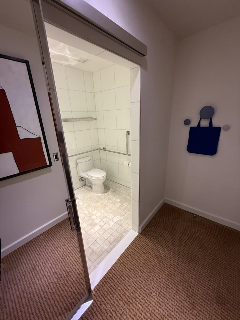 Accessible bathroom doorway at Hotel Park Ave seen from the bedroom hallway. Sliding door pulled back into the wall. Flush threshold between bedroom carpet and bathroom tile. Toilet with side grab rail visible inside. White wall tile and small-format floor tile.