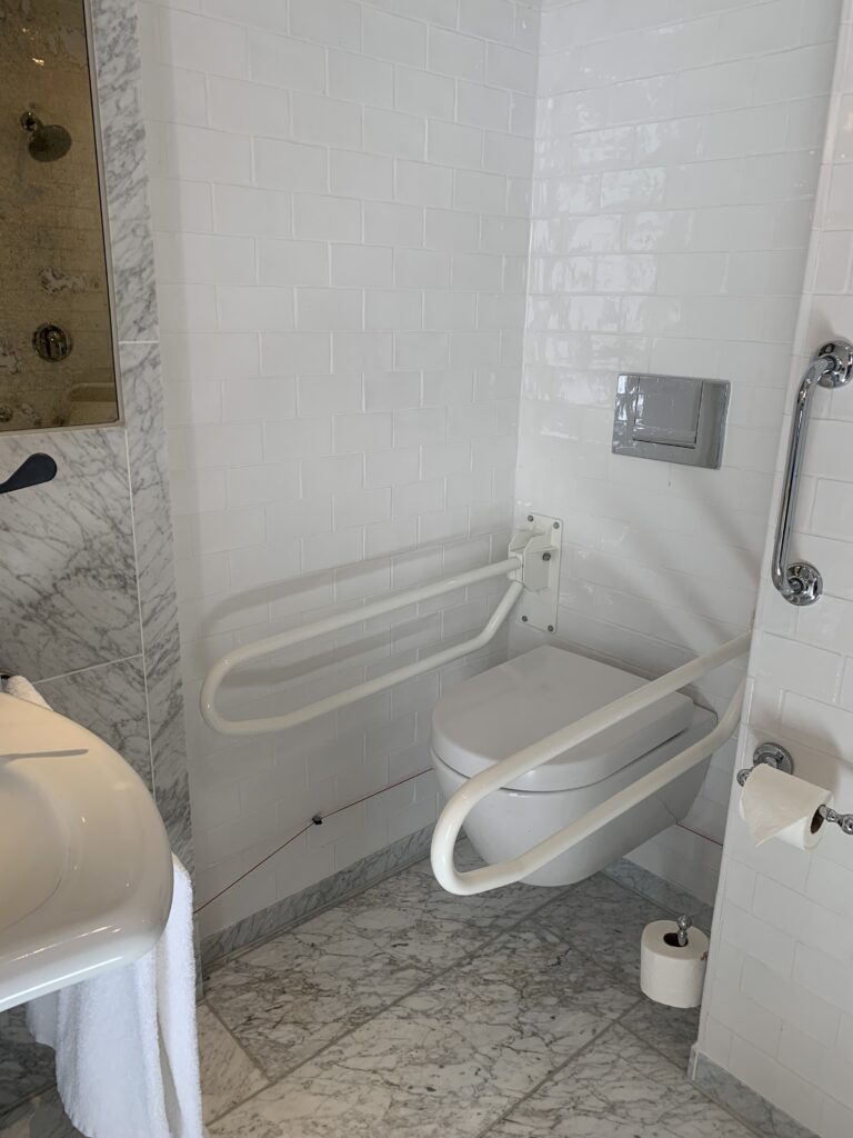 Wall-hung toilet in a Pulitzer accessible bathroom with hinged drop-down grab rails on both sides, a wall-mounted basin to the left, a flush plate and toilet roll holder, and white subway-tiled walls above a marble floor.