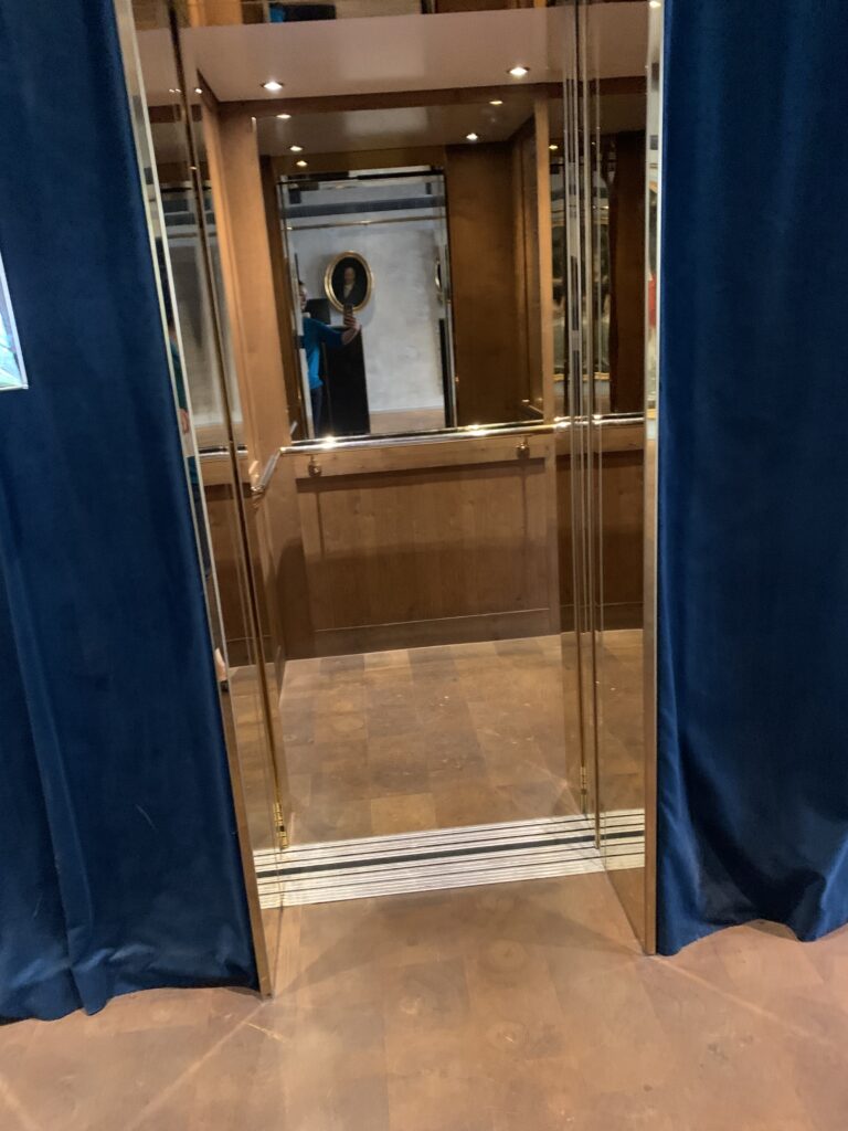 Guest lift at Pulitzer Amsterdam. Brass-panelled cab with mirrored walls, wood lower panelling, handrail, doors open at a level threshold.
