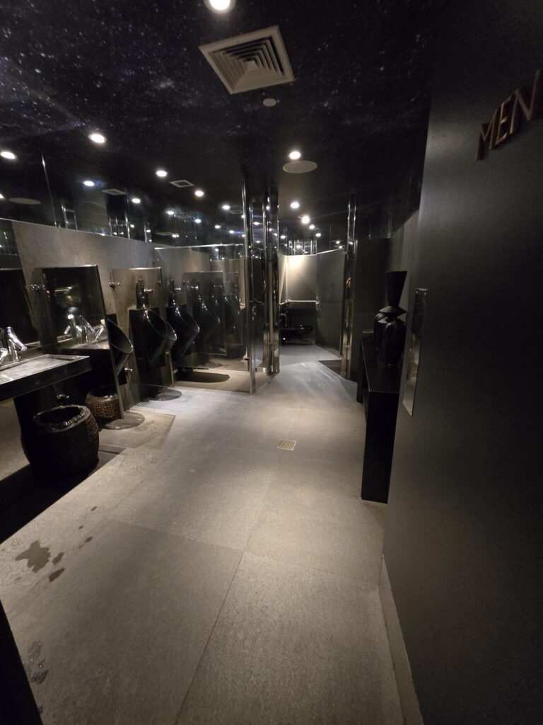 Public washroom at Hotel Park Ave NYC. Mirrored ceiling, run of urinals and sinks along one wall, cubicles opposite, with an accessible cubicle at the end.