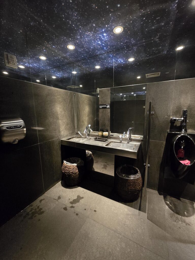 Washbasin area in the public washroom at Hotel Park Ave NYC. Double trough sink on a counter with open clearance beneath, mirror above, hand dryer on the side wall.