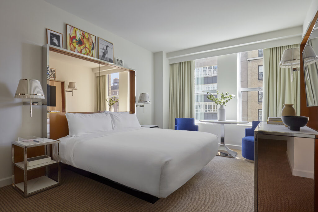 Premier Studio King at Hotel Park Ave NYC. King bed against a mirrored headboard wall with framed artwork above, twin pleated wall lights, two blue velvet armchairs and a round table at the window, mirrored desk and nightstands, city view.