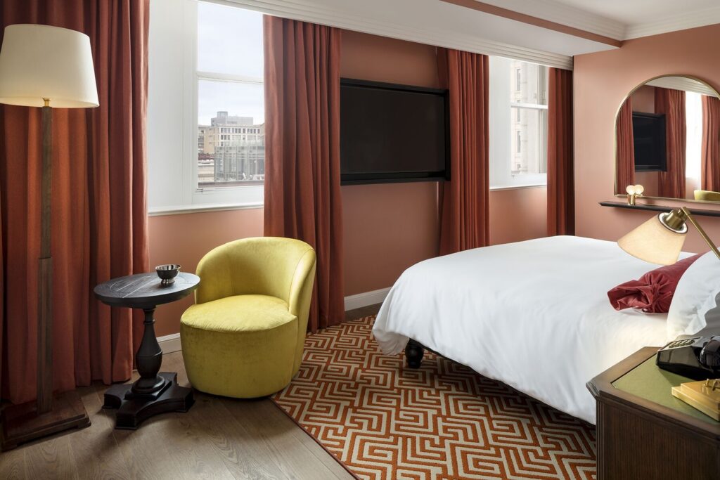 Riggs Desk JPG. And the alt text is Guest room at Riggs Washington DC. Terracotta walls, bed with a city view through the window, yellow tub chair and round side table, patterned rug, wall-mounted television, floor lamp.