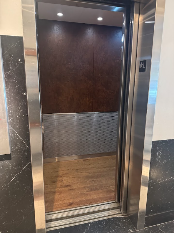 Guest lift interior at Lyle Washington DC. Wood-panelled cab with a textured metal lower wall and wood floor. Doors open at floor level with a level threshold into the cab.