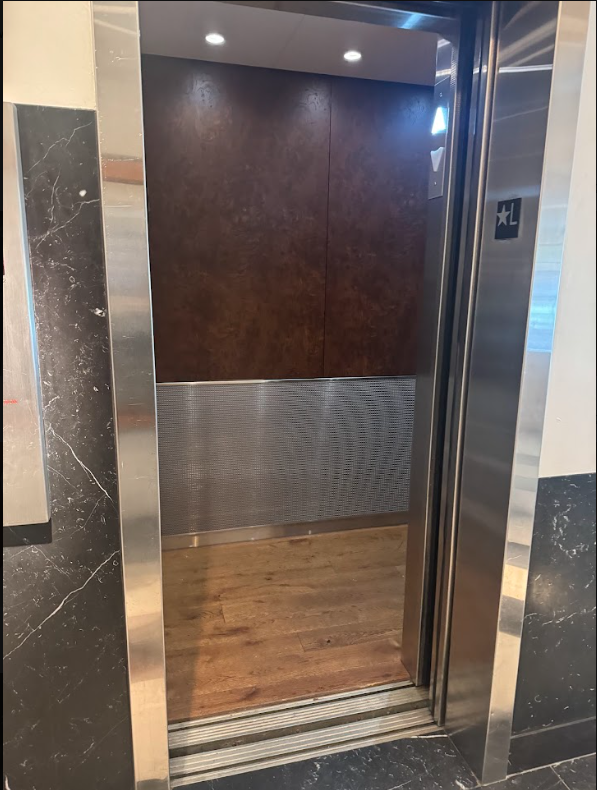 Guest lift interior at Lyle Washington DC. Wood-panelled cab with a textured metal lower wall and wood floor. Doors open at floor level with a level threshold into the cab.